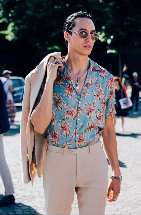 A bold tropical wedding guest look with a blue printed shirt a tan linen suit and layered necklaces is cool and chic