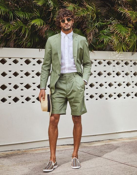 A chic tropical wedding gust look with a green suit that includes shorts a white button down and printed slipons plus a straw hat