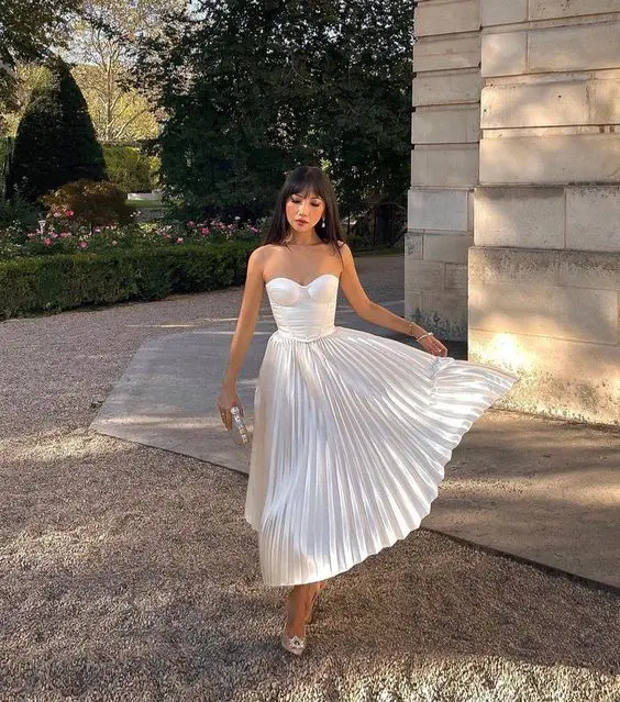 an exquisite white strapless midi dress with a corset and a pleated skirt nude shoes and a small bag